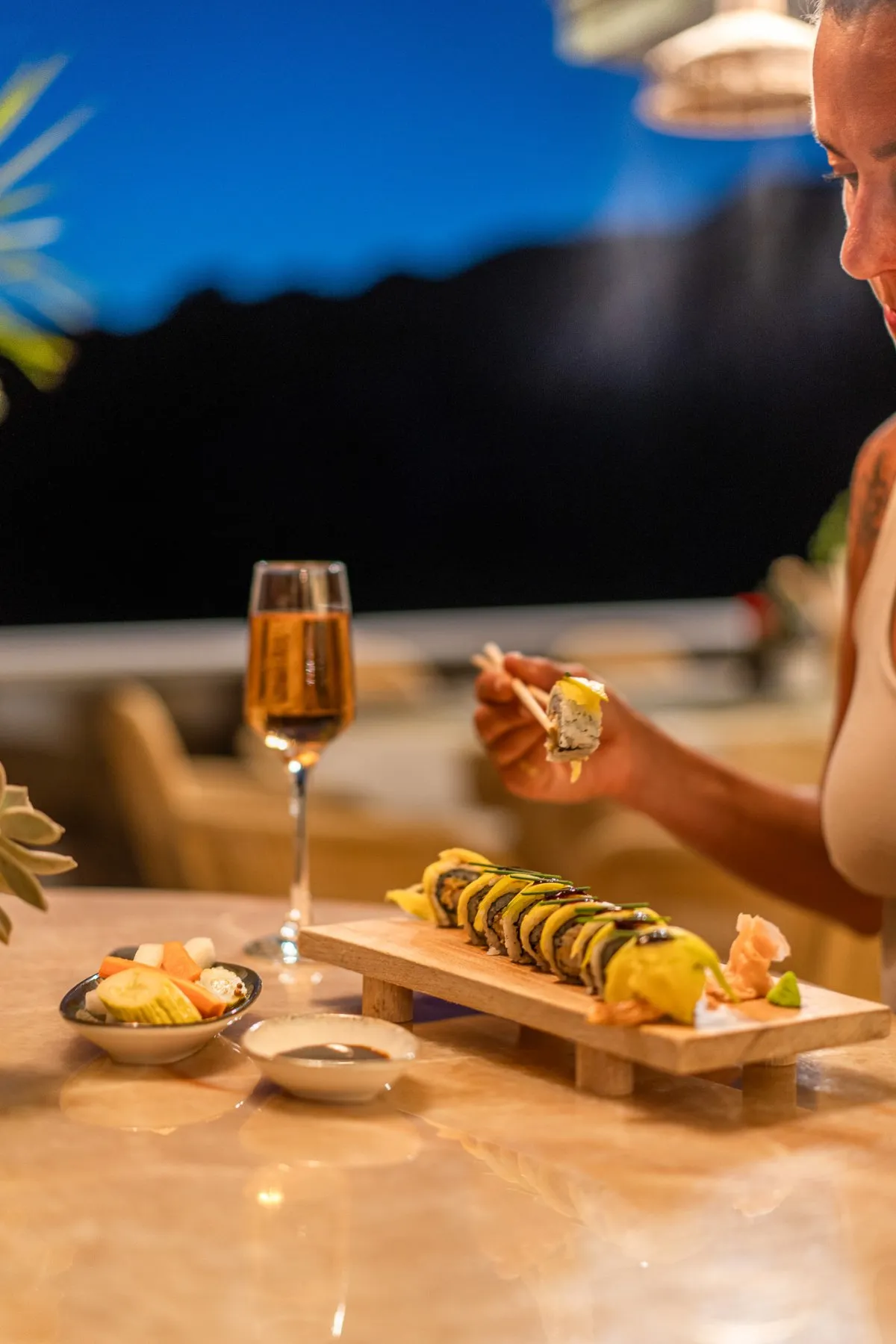 Mavi saatte tahta altlıkta bir sushi rolü ve bir kadeh roze; bir misafir çubuklarla bir parça alıyor.