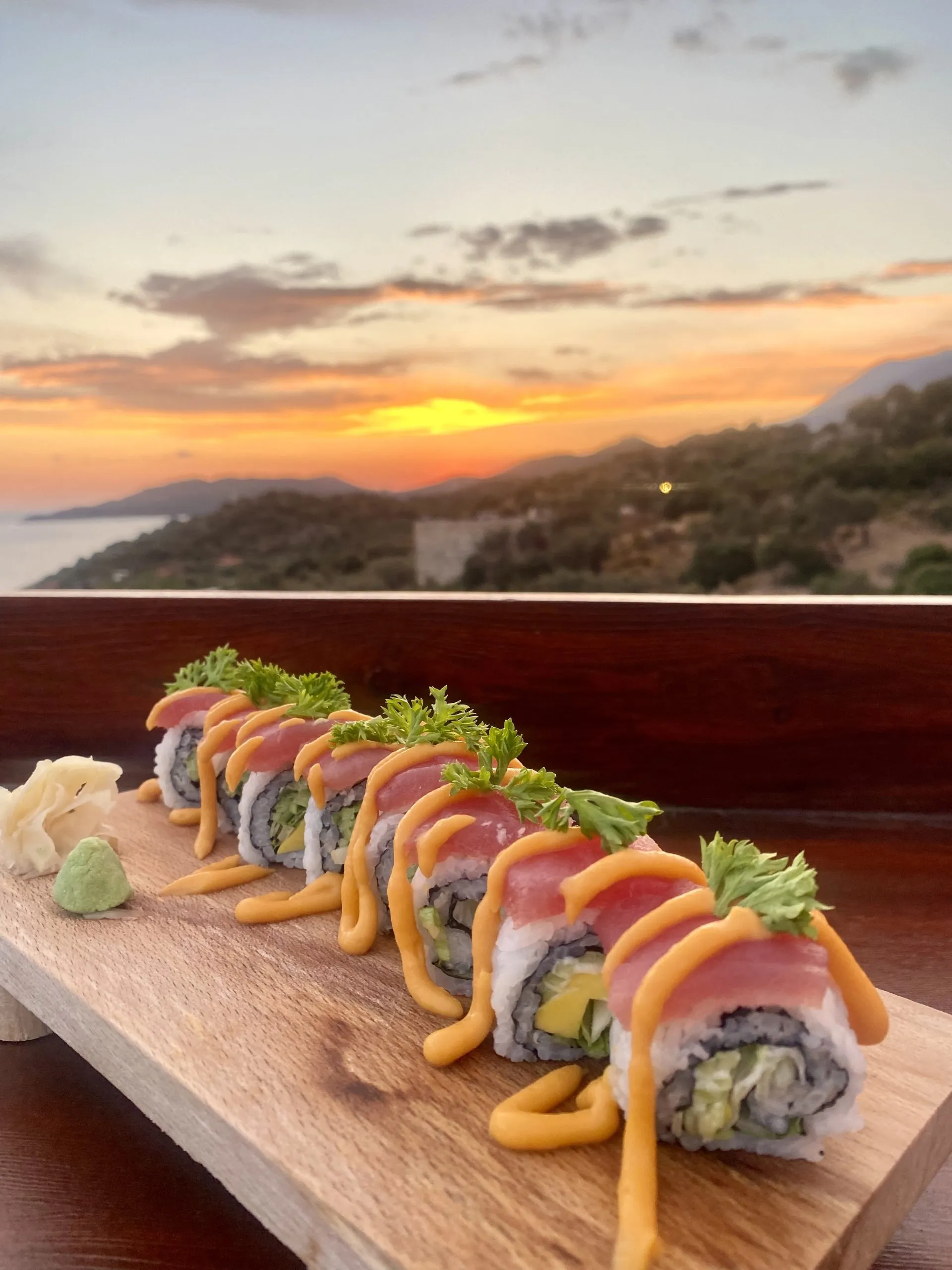 Gün batımında terasın tahta korkuluğunda, altlıkta üstü balıklı ve turuncu soslu bir sushi rolü; arkada güneş Kaş'ın tepelerinin ardına iniyor, solda deniz.